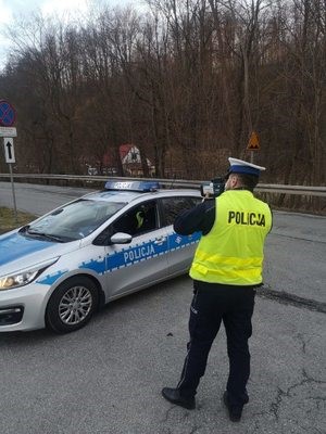 policjant stoi przy radiowozie na drodze i nadzoruje ruch, w ręku trzyma miernik prędkości
