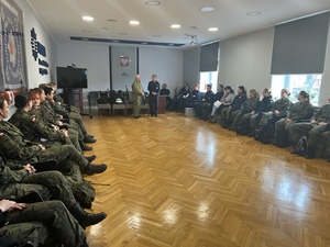 grupa uczniów w mundurach ćwiczebnych siedzi na krzesłach w sali sztabowej, na środku stoi mężczyzna w mundurze polowym oraz policjantka