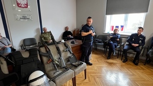 grupa uczniów w mundurach ćwiczebnych siedzi na krzesłach w sali sztabowej, na środku stoi policjant
