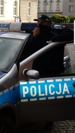 Policjant w mundurze stoi przy radiowozie trzyma w ręku mikrofon i przekazuje komunikat dla mieszkańców. W tle widać budynki rynku w porze dziennej.