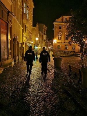 Policjanci sprawdzają czy mieszkańcy dostosowują się do zaleceń. Dwóch policjantów idzie przez oświetlone latarniami ulice miasta wieczorem