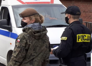Policjant umundurowany stoi przy karetce pogotowia i trzyma w ręku kartkę papieru,a z jego lewej strony stoi kobieta żołnierz.