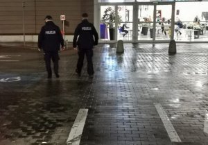 Wieczorem dwóch umundurowanych policjantów idzie w kierunku galerii handlowej. Z prawej i lewej  ich strony widać zaparkowane samochody osobowe.