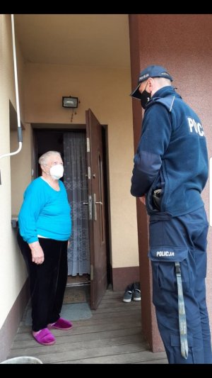 Policjant w mundurze i maseczce na twarzy stoi przy otwartych drzwiach do domu obok niego stoi starsza kobieta ubrana w sweter, spodnie oraz kapcie. Kobieta  na twarzy ma maseczkę.