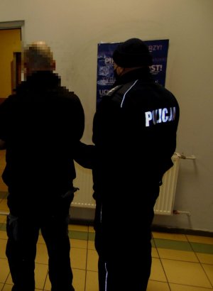 policjant w mundurze stoi z zatrzymanym meżczyną. Osoby są na zdjęciu odwrócone tyłem za nimi znajduje się ściana pomieszczenia w którym przebywają.