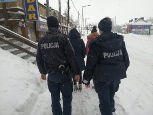 policjant w mundurze oraz dwie kobiety idą w kierunku budynku. Dookoła jest śnieg. Osoby są sfotografowane od tyłu.W oddali widać obiekty handlowo-usługowe.Zdjęcie wykonane w porze dziennej