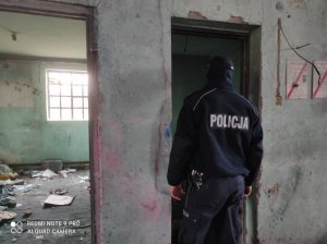 policjant umundurowany stoi w pomieszczeniu pustostanu, gdzie ściany są brudne i porysowane a na podłodze leża śmieci