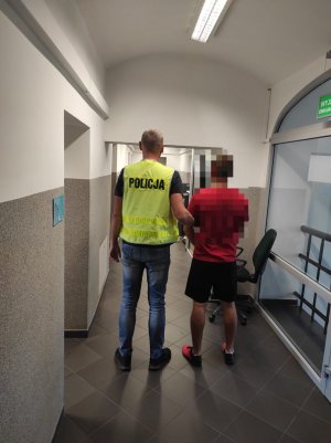 policjant prowadzi zatrzymanego mężczyznę , osoby są pomieszczeniu służbowym