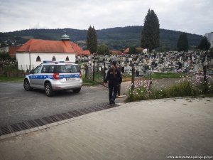 policjantka stoi przy wejściu na cmentarz , obok zaparkowany radiowóz policyjny