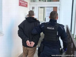 policjant prowadzi zatrzymanego mężczyznę w kierunku drzwi na ścianie tabliczka z napisem Prokuratura Rejonowa