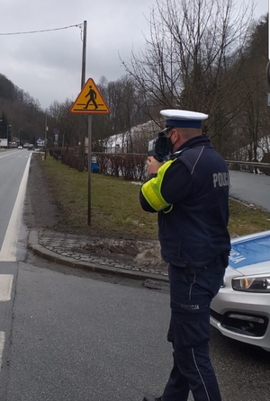 policjant trzyma w ręku miernik prędkości i stoi na drodze przy radiowozie