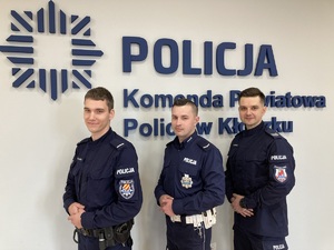 TRZECH POLICJANTÓW STOI PRZY ŚCIANIE Z NAPISEM kOMENDA pOWIATOWA POLIJI W kŁODKZU