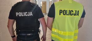 dwóch policjantów stoi tyłem , maja na sobie kamizelki policyjne