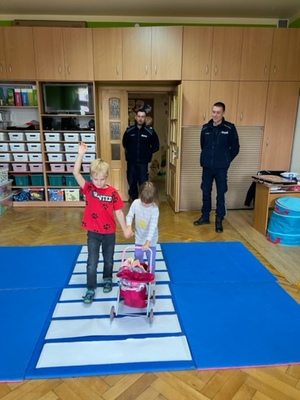 dwóch policjantów stoi za dziećmi  w sali przedszkola.Dzieci ida przez matę z pasami