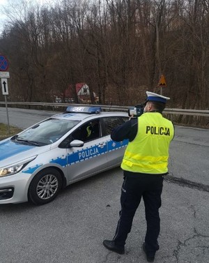 policjant stoi na drodze z miernikiem prędkości w ręku obok zaparkowany radiowóz