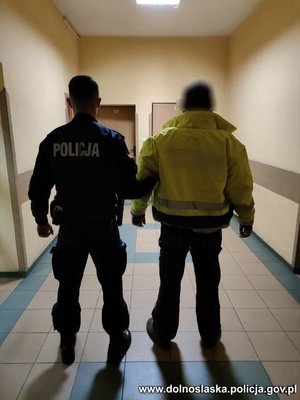 policjant w pomieszczeniu prowadzi zatrzymanego mężczyznę, który ma kajdanki na rękach