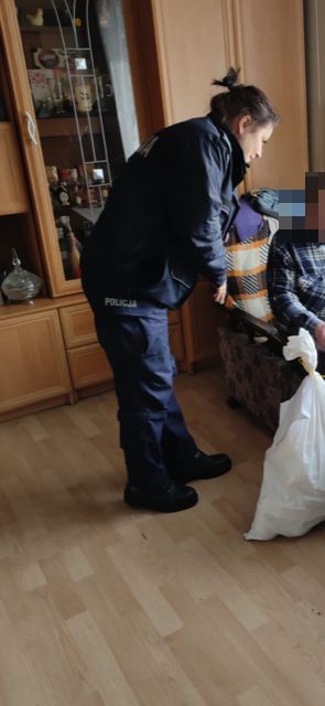 policjantka stoi  w pokoju osoby starszej i obok leży worek