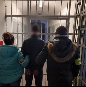 Policjant i policjantka w ubraniu cywilnym z opaskami na ręku z napisem policja prowadzą zatrzymanego mężczyznę w kierunku krat. Zdjęcie wykonane w pomieszczeniu.