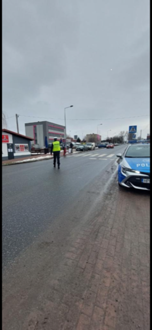 policjant stoi przed przejściem, widać pieszych , na poboczu zaparkowany radiowóz