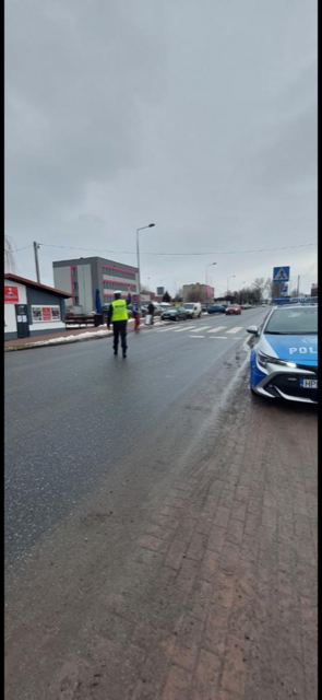 policjant stoi przy przejściu dla pieszych, widać zaparkowany radiowóz policyjny