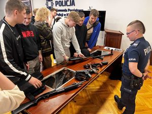 Obok biurka stoi policjant, na biurku leży broń, przed biurkiem stoi młodzież