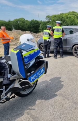 policjanci stoją przy samochodzie , widać fragment motocyklu policyjnego oraz przedstawiciela służb drogowych