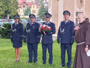 Zdjęcie z uroczystości święta Policji, złożenie wiązanki przy obelisku w ogrodach Ojców Franciszkanów w Kłodzku -grupa policjantów oraz duchowny