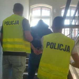 Policjanci w kamizelkach z napisem Policja prowadzą po schodach zatrzymanego mężczyznę, który ma kajdanki założone z tyłu na rękach