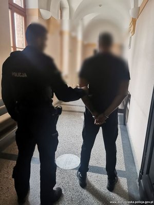 policjant prowadzi zatrzymanego mężczyznę , który ma założone z tyłu kajdanki , zdjęcie wykonane w pomieszczeniu