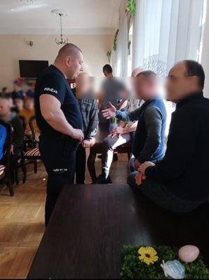 Policjant stoi przy mężczyznach w pomieszczeniu i rozmawia, w tle widać osoby siedzące na krzesłach