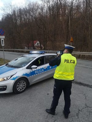 policjant trzyma w rękach miernik prędkości , stoi przy radiowozie policyjnym na poboczu drogi, widać ulice, a w tle widać drzewa