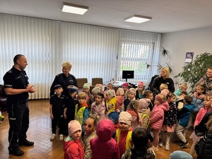 policjant i policjantka stoją obok   grupy dzieci  w pomieszczeniu policji