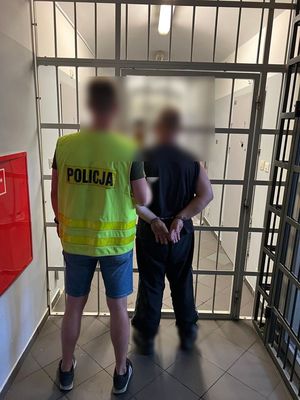 policjant mający na sobie kamizelkę z napisem Policja prowadzi zatrzymanego mężczyznę , który ma kajdanki z tyłu na rękach