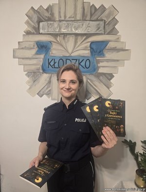 policjantka stoi przy ścianie z logo policji kłodzkiej i trzyma w ręku książeczki