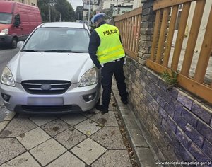 policjant próbuje przejść przy zaparkowanym na chodniku samochodzie osobowym, widać c fragment busa na zdjęciu