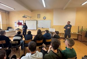 policjantka stoi pzred grupą siedzacych na krzesłach młodych ludzi-uczniów w klasie