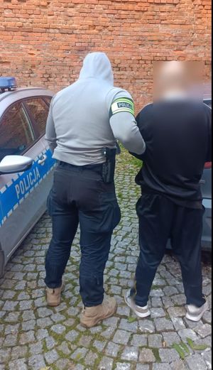 policjant prowadzi zatrzymanego mężczyznę. który ma ręce do tyłu i kajdanki na nich. widać fragment radiowozu policyjnego