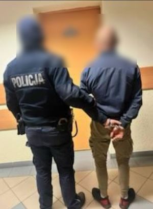 policjant prowadzi  zatrzymanego mężczyznę, który na rękach z tyłu ma kajdanki