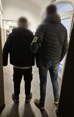 policjant z opaską na ręce na której jest napis POLICJA  prowadzi zatrzymanego mężczyznę w pomieszczeniu służbowym - korytarz