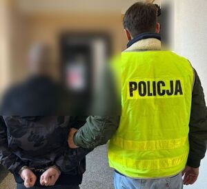 policjant prowadzi w pomieszczeniu zatrzymanego mężczyznę , który ma rękach z tyłu kajdanki