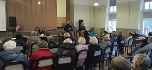 policjant stoi obo0k kobiety a przed  nim w sali siedzą osoby na krzesłach