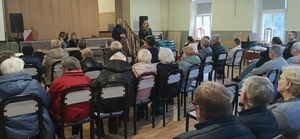 policjant stoi obo0k kobiety a przed  nim w sali siedzą osoby na krzesłach