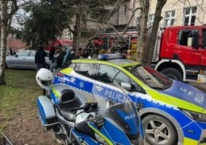 radiowóz i inne pojazdy służb stoją przy budynku w tle widać młodzież i mundurowych