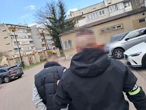 Policjant z opaską na ręce prowadzi zatrzymanego mężczyznę.