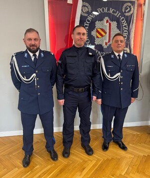 trzech policjantów  stoi w sali sztabowej przy sztandarze, który jest na ścianie w sali sztabowej