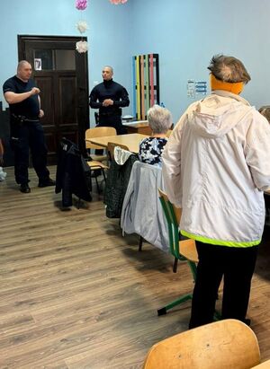dwóch policjantów  w sali stoi przed osobami, które siedzą na krzesełkach, a jedna osoba stoi  - spotkanie