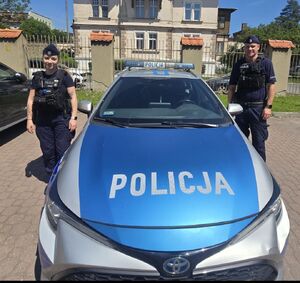 policjant i policjantka stoją przy radiowozie policyjnym w tle widać budynek