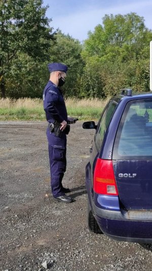 Zadanie patrolujących policjantów jak mieszkańcy respektują obowiązujące nakazy. Policjant stoi przy samochodzie osobowym na którym w tylnej części jest napis Golf i prowadzi kontrole. W tle widać krzaki i trawę