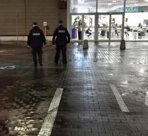 Wieczorem dwóch umundurowanych policjantów  idzie w kierunku galerii handlowej, W tle widać za szybą osoby znajdujące się w galerii.