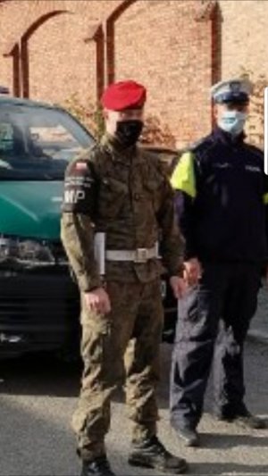 Policjant Wydziału Ruchu Drogowego z białą czapką na głowie  oraz żołnierz Żandarmerii Wojskowej z czerwonym beretem na głowie stoją przy samochodzie oznakowanym Żandarmerii Wojskowej . Mundurowi mają na twarzy maseczki. W tle widać mur.